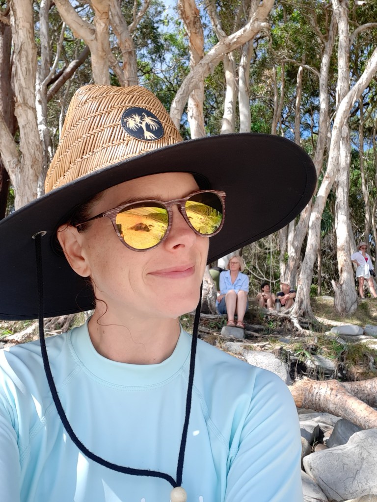 Person wearing a wide-brimmed hat and sunglasses, smiling in a natural outdoor setting with trees in the background.