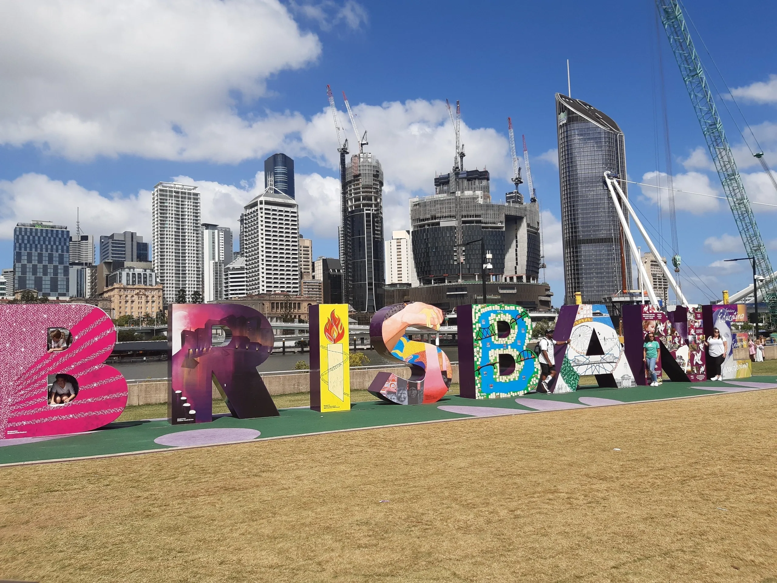 Colorful 'BRISBANE' letters displayed in a park, with a skyline of modern buildings and construction cranes in the background under a blue sky.